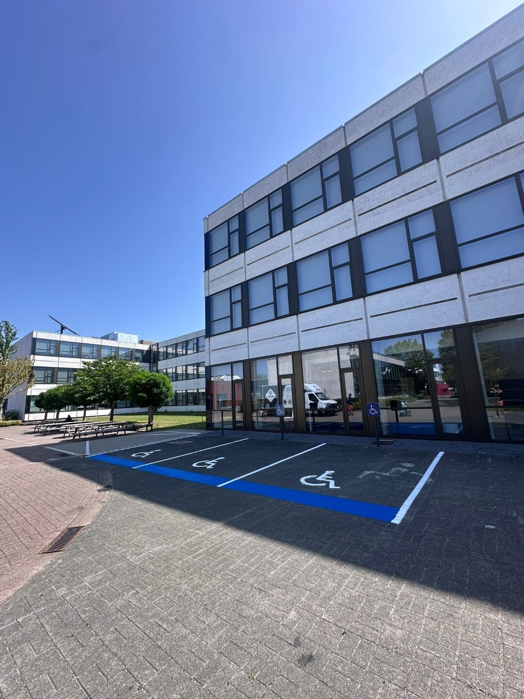 Een modern kantoorgebouw met witte gevelpanelen en grote ramen, gelegen aan een plein met meerdere blauwe gehandicaptenparkeerplaatsen voor de ingang; de lucht is helderblauw en er staan bomen op de achtergrond.